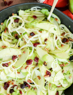 A bowl of fresh Apple Fennel Salad with thinly sliced green apples, celery, onions, dried cranberries, and chopped pecans. Two green apples and an orange napkin are nearby, with scattered pecans and cranberries on the table.