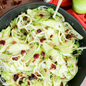 A bowl of fresh Apple Fennel Salad with thinly sliced green apples, celery, onions, dried cranberries, and chopped pecans. Two green apples and an orange napkin are nearby, with scattered pecans and cranberries on the table.