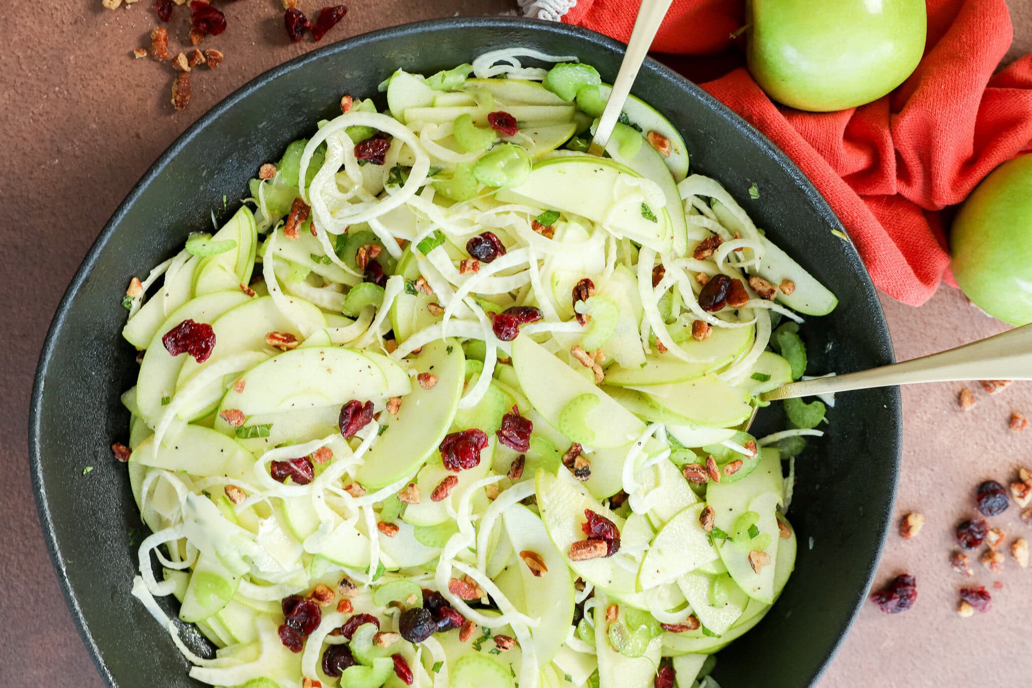 A bowl of fresh Apple Fennel Salad with thinly sliced green apples, celery, onions, dried cranberries, and chopped pecans. Two green apples and an orange napkin are nearby, with scattered pecans and cranberries on the table.