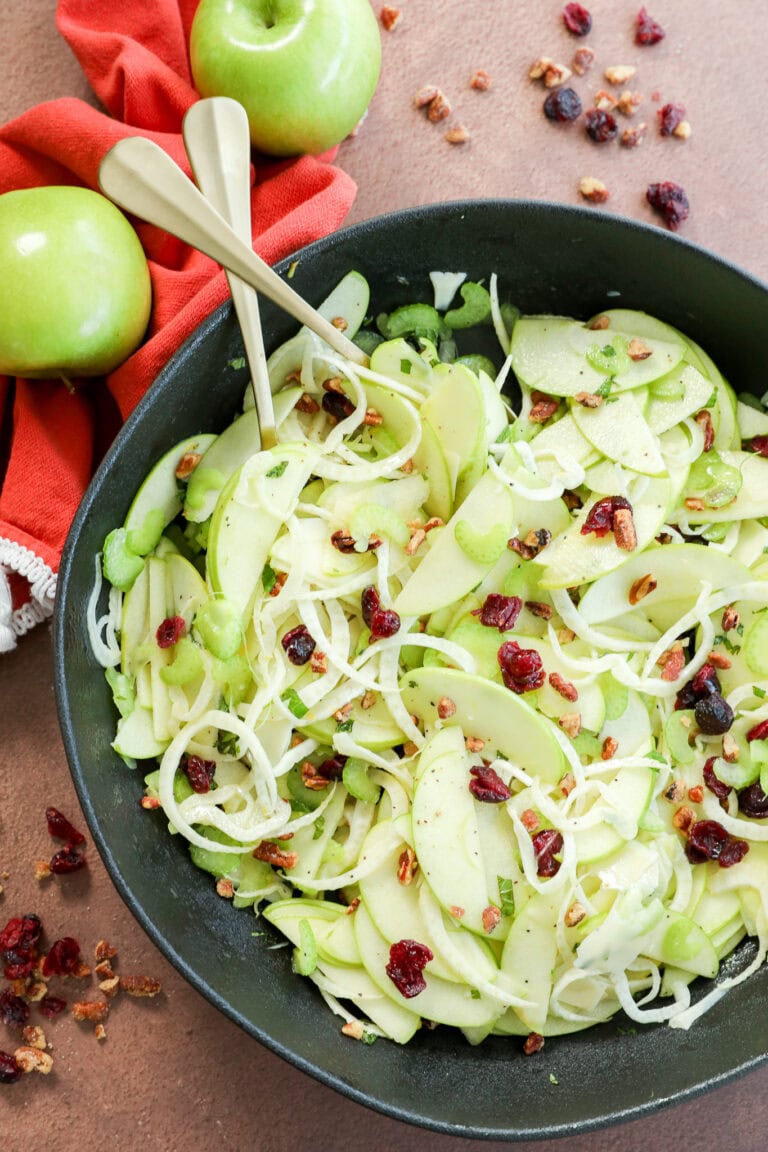 A bowl of fresh Apple Fennel Salad made with thinly sliced green apples, shredded cabbage, celery, dried cranberries, and chopped nuts. Two serving utensils rest in the bowl, with whole green apples and a red napkin beside it.