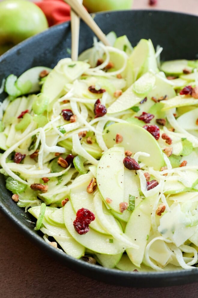A close-up of an Apple Fennel Salad with thinly sliced green apples, fennel, dried cranberries, chopped nuts, and herbs in a black bowl, with wooden chopsticks resting inside.