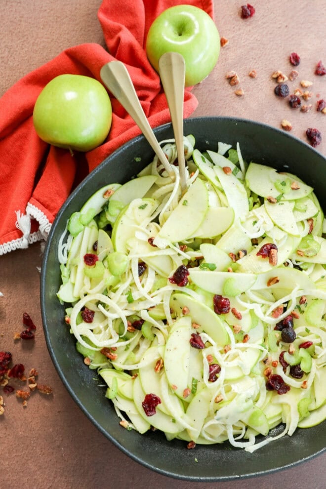 A black bowl filled with an Apple Fennel Salad of thinly sliced green apples, fennel, celery, dried cranberries, and nuts. Two serving utensils rest in the bowl. A red cloth and two whole green apples are beside the bowl.