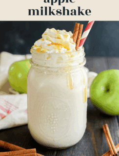 A mason jar filled with apple milkshake topped with whipped cream, caramel drizzle, and two cinnamon sticks, surrounded by green apples and a striped straw on a dark wooden table.
