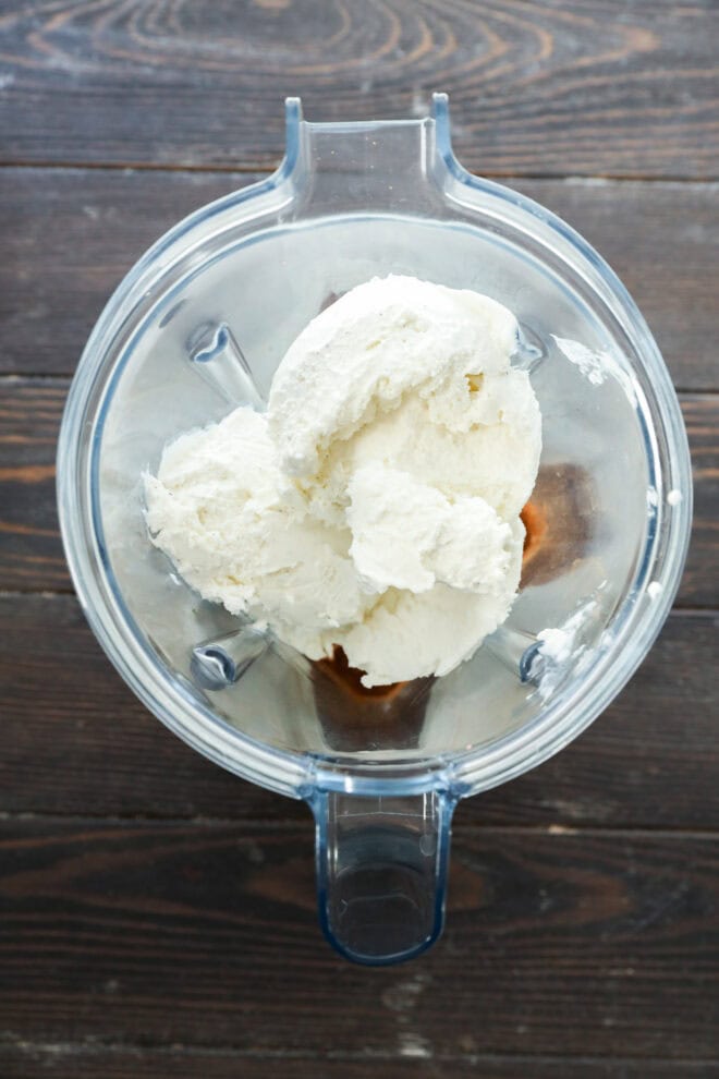 A top-down view of a blender containing scoops of vanilla ice cream and a few brown-colored ingredients, sitting on a dark wooden surface.