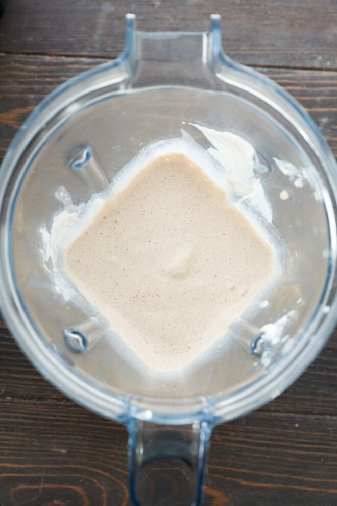 Overhead view of a blender containing a creamy, light beige mixture with some residue on the sides, sitting on a dark wooden surface.