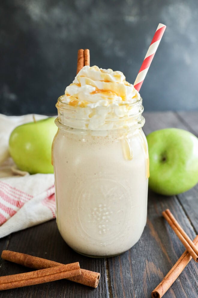 A mason jar filled with a creamy apple milkshake topped with whipped cream, caramel drizzle, and cinnamon sticks, with a red-and-white striped straw. Green apples and cinnamon sticks are on a rustic wooden table in the background.