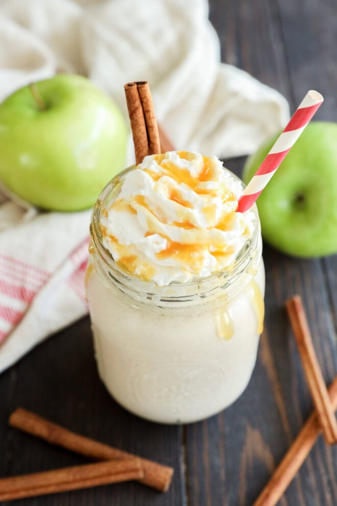 A mason jar filled with a creamy drink topped with whipped cream, caramel drizzle, and two cinnamon sticks, with a red and white striped straw. Green apples and cinnamon sticks are nearby on a wooden surface.