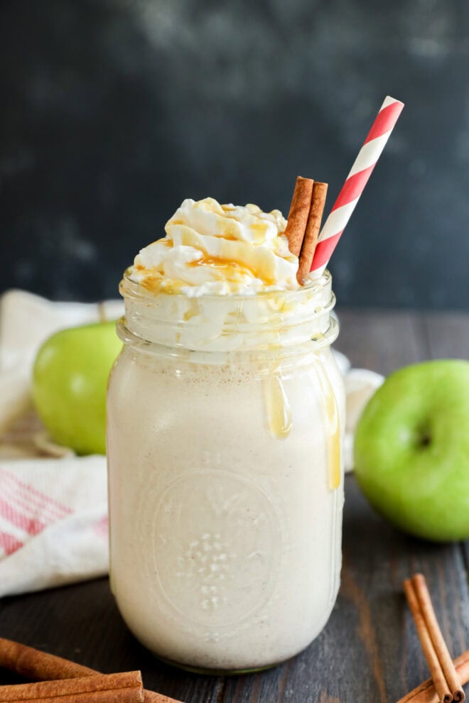 A mason jar filled with a creamy milkshake, topped with whipped cream, caramel drizzle, two cinnamon sticks, and a red-striped straw. Green apples and cinnamon sticks are in the background on a dark wooden surface.