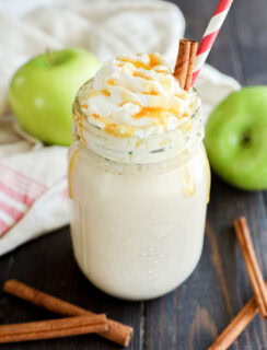 A creamy smoothie topped with whipped cream and caramel sauce in a mason jar, garnished with cinnamon sticks and a red-striped straw. Green apples and more cinnamon sticks are on the wooden table.