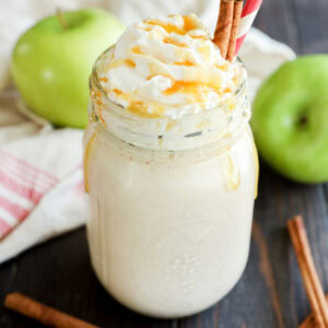 A creamy smoothie topped with whipped cream and caramel sauce in a mason jar, garnished with cinnamon sticks and a red-striped straw. Green apples and more cinnamon sticks are on the wooden table.