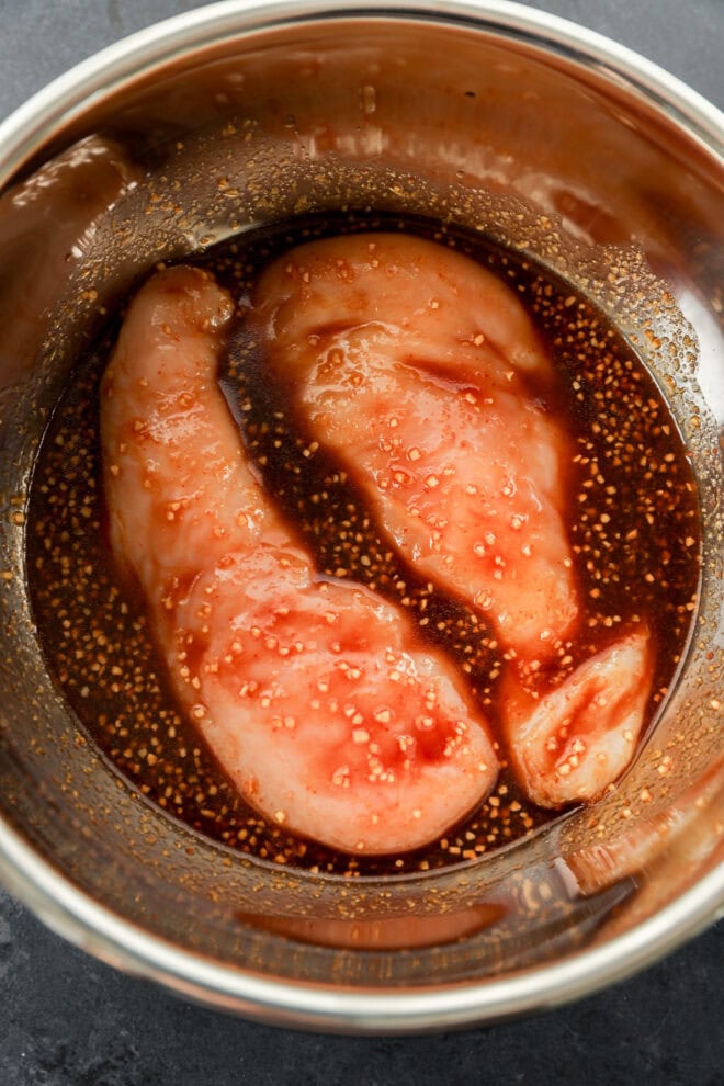 Two raw chicken breasts marinate in a stainless steel bowl with a dark Asian Chicken Marinade packed with aromatic spices.