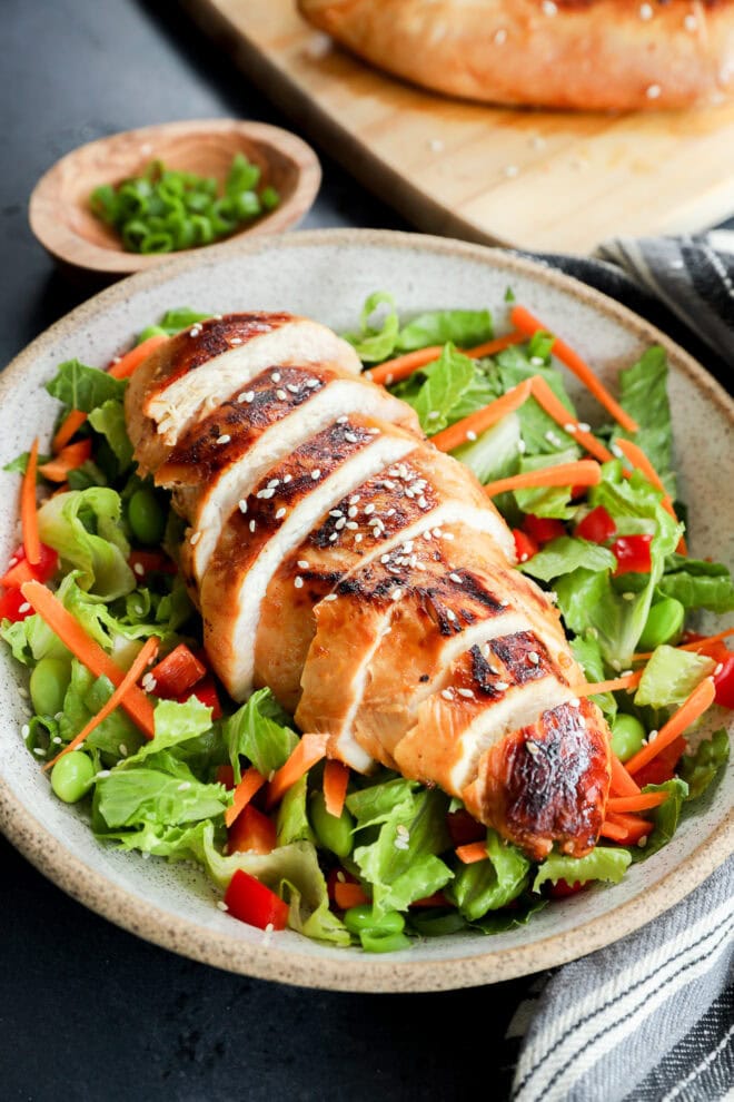 A plate with sliced grilled chicken breast, marinated in an Asian Chicken Marinade, served over mixed greens, shredded carrots, red bell peppers, and edamame, garnished with sesame seeds. A wooden bowl with chopped green onions sits in the background.