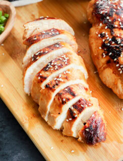 Sliced grilled chicken breast, topped with sesame seeds, rests on a wooden cutting board, with part of another chicken breast and a small bowl of green onions nearby.