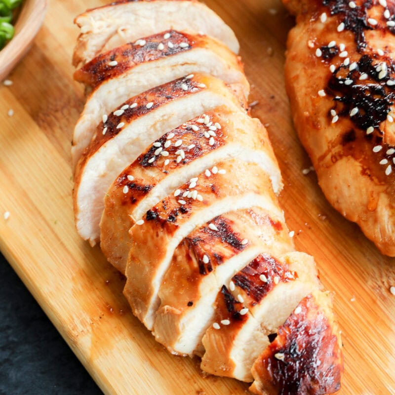 Sliced grilled chicken breast, topped with sesame seeds, rests on a wooden cutting board, with part of another chicken breast and a small bowl of green onions nearby.