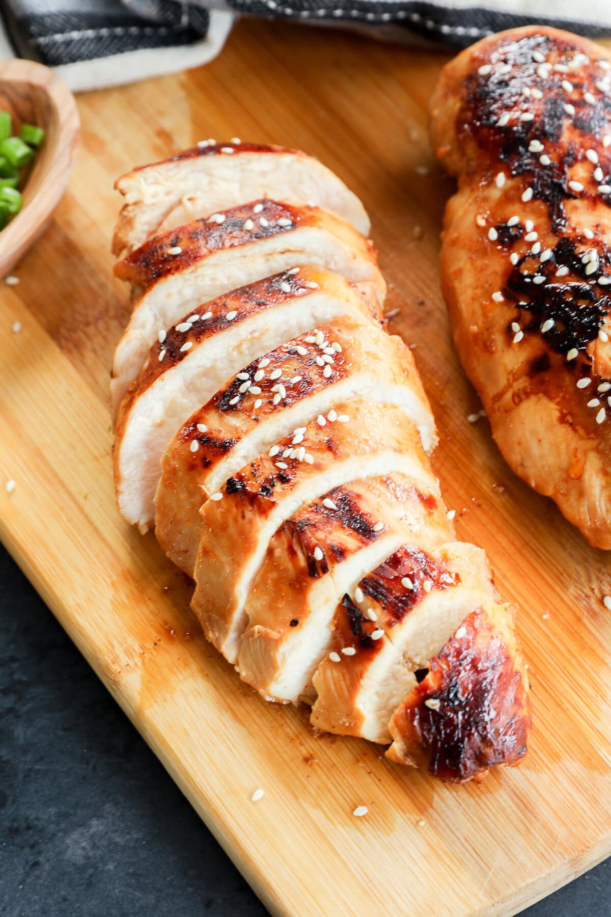 Sliced grilled chicken breast, topped with sesame seeds, rests on a wooden cutting board, with part of another chicken breast and a small bowl of green onions nearby.