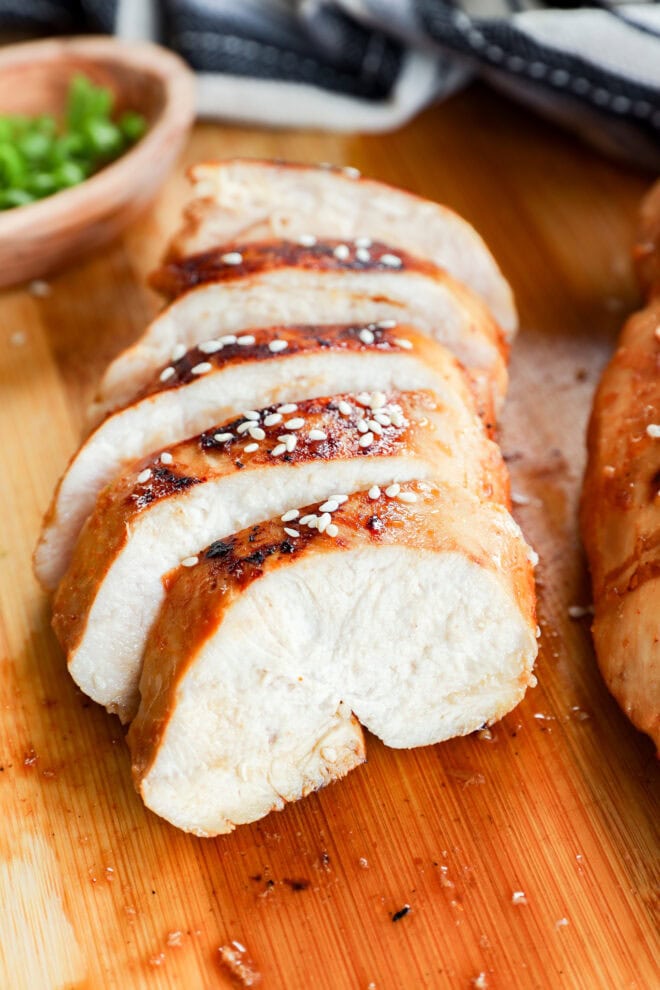 Sliced cooked chicken breast, marinated in a savory Asian Chicken Marinade and sprinkled with sesame seeds, is arranged neatly on a wooden cutting board with a small bowl of chopped green onions in the background.