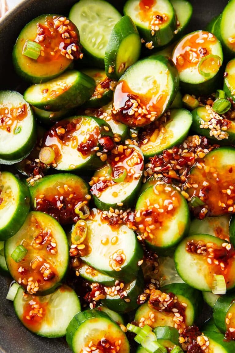 Close-up of sliced cucumbers topped with a spicy chili oil sauce, sesame seeds, and chopped green onions. The dish appears fresh, flavorful, and has a glossy, vibrant appearance.
