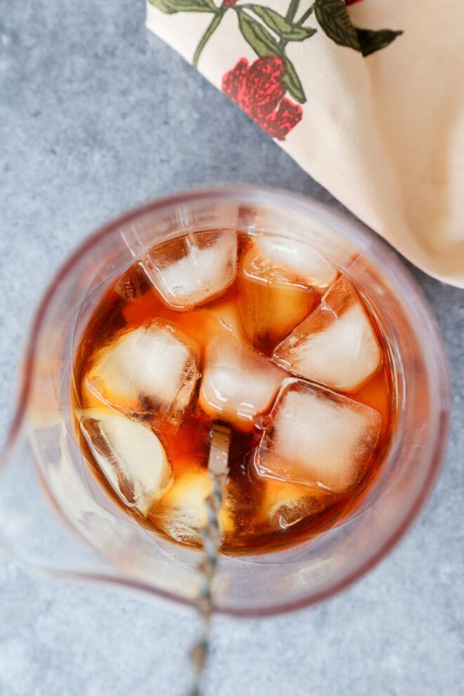 Overhead view of a glass filled with amber liquid and several ice cubes, likely a Black Manhattan, with a metal mixing spoon inside. A beige napkin with a red floral pattern is partially visible in the corner.