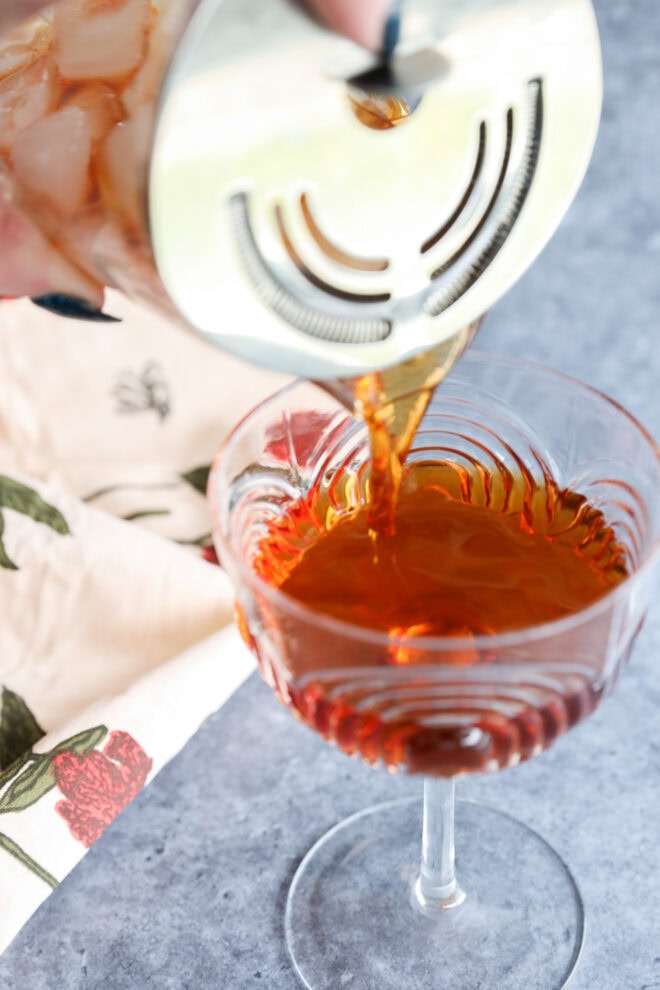 A Black Manhattan is being poured through a strainer into an elegant glass, with a floral-patterned napkin nearby on a gray surface.