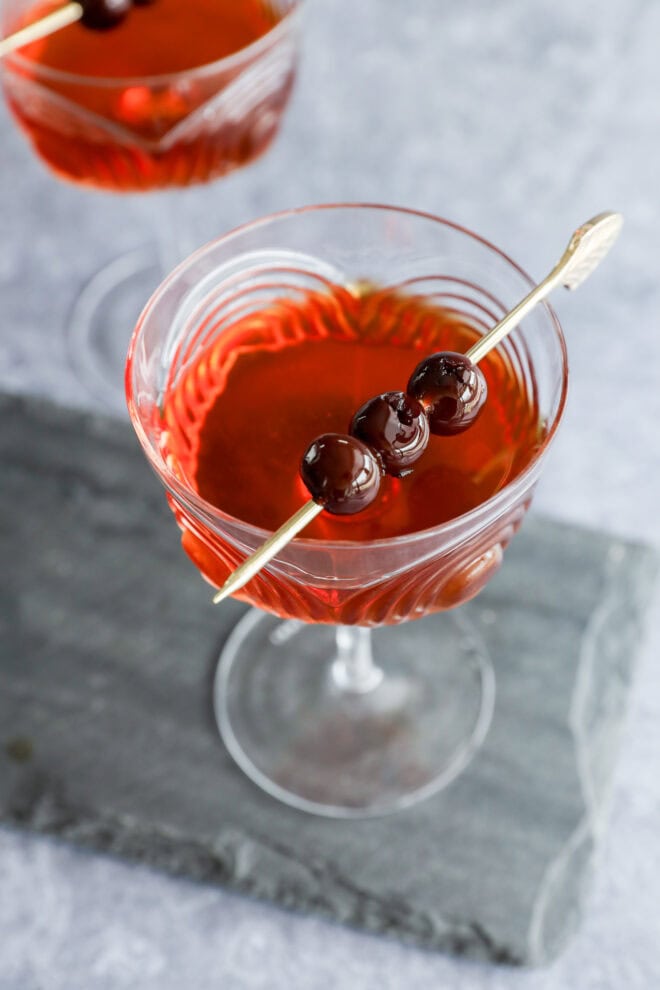 A Black Manhattan cocktail in a clear glass, garnished with three dark cherries on a cocktail pick, rests on a gray stone slab, with another similar drink partially visible in the background.