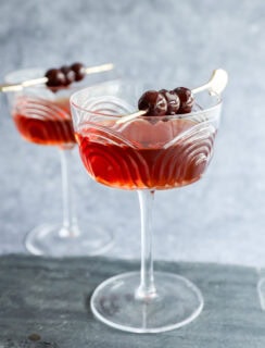 Two elegant coupe glasses filled with a Black Manhattan cocktail, each garnished with three dark cherries on a cocktail pick, set on a slate surface with a gray background.