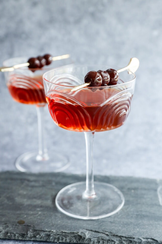 Two elegant coupe glasses filled with a Black Manhattan cocktail, each garnished with three dark cherries on a cocktail pick, set on a slate surface with a gray background.