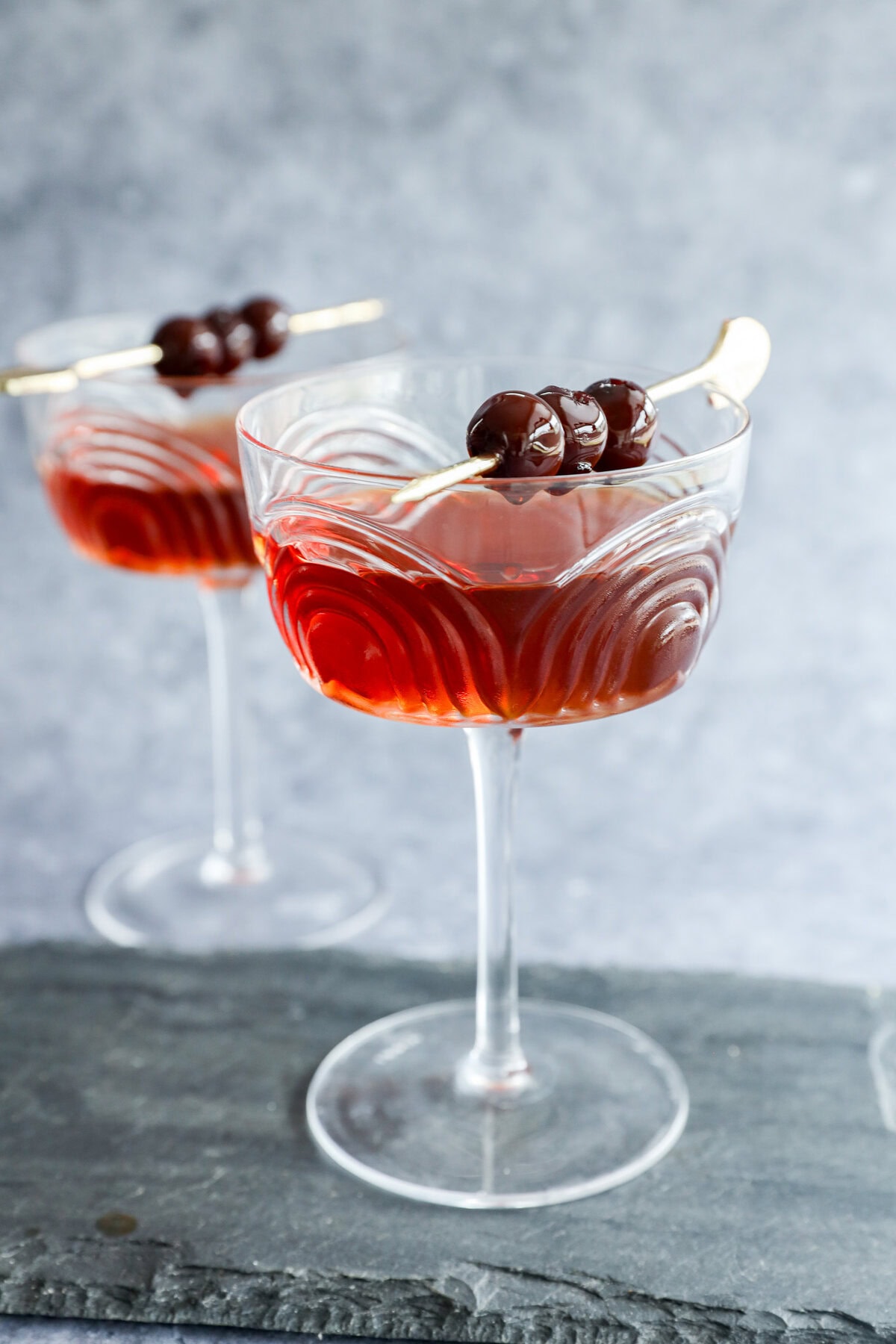 Two elegant coupe glasses filled with a Black Manhattan cocktail, each garnished with three dark cherries on a cocktail pick, set on a slate surface with a gray background.
