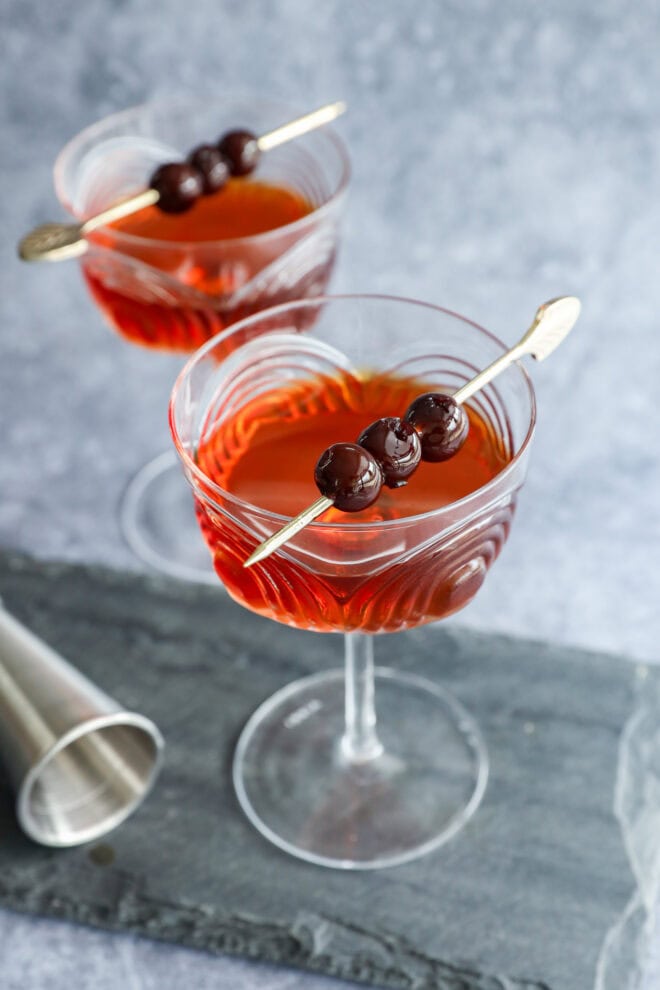 Two crystal cocktail glasses filled with a reddish-brown Black Manhattan, each garnished with three dark cherries on a cocktail pick. A metal jigger sits nearby on a gray slate surface.