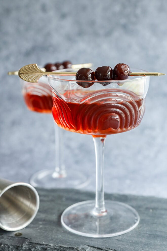 Two elegant cocktail glasses filled with a red Black Manhattan, each garnished with three dark cherries on a gold skewer. The wavy-patterned glasses rest on a slate surface, with a cocktail shaker nearby.
