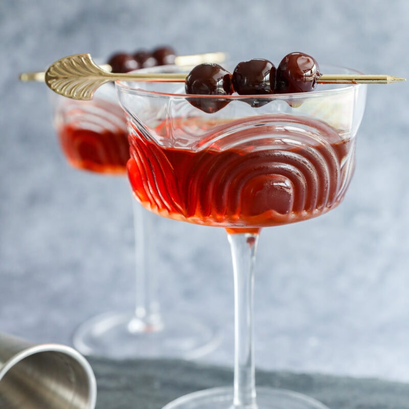 Two elegant cocktail glasses filled with a red Black Manhattan, each garnished with three dark cherries on a gold skewer. The wavy-patterned glasses rest on a slate surface, with a cocktail shaker nearby.