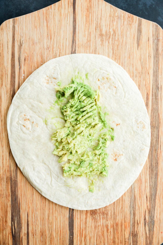 A flour tortilla on a wooden board with smashed avocado spread in the center.