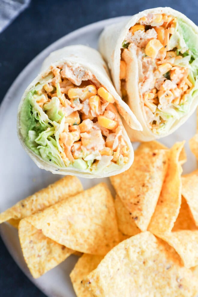 A plate with a wrap cut in half, filled with lettuce, corn, chicken, and sauce, served alongside tortilla chips.