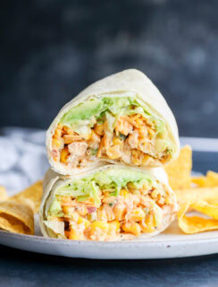 Two halves of a wrap filled with diced chicken, lettuce, and sauce are stacked on a plate with tortilla chips on the side, against a dark blurred background.