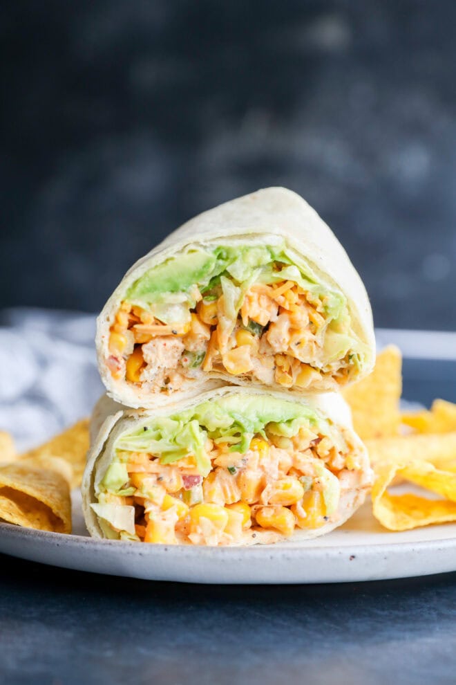 Two halves of a wrap filled with diced chicken, lettuce, and sauce are stacked on a plate with tortilla chips on the side, against a dark blurred background.