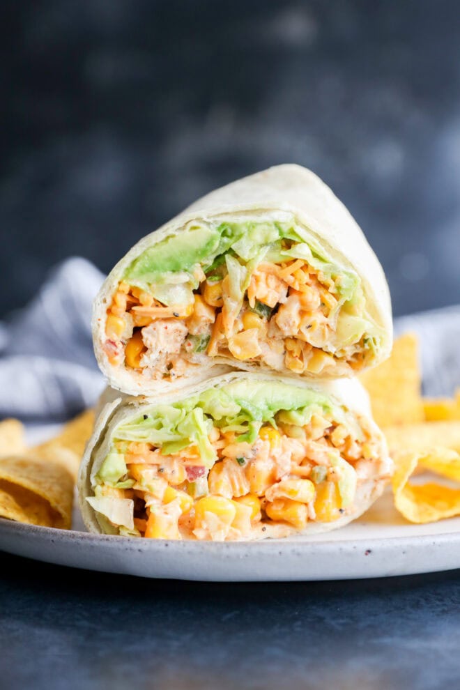A close-up of a wrap cut in half, filled with lettuce, corn, diced vegetables, and a creamy filling, served on a plate with tortilla chips.