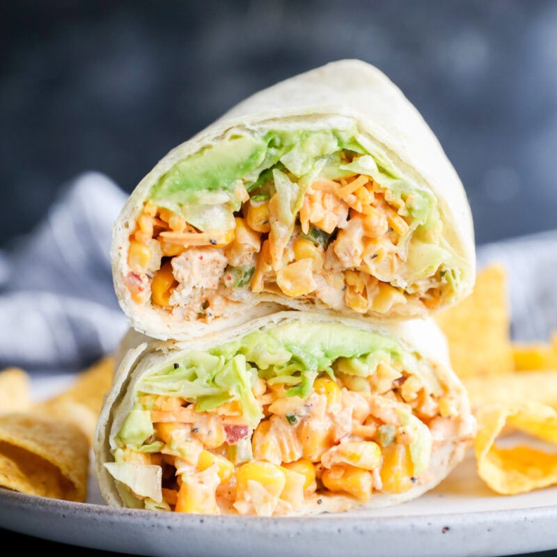 A close-up of a wrap cut in half, filled with lettuce, corn, diced vegetables, and a creamy filling, served on a plate with tortilla chips.