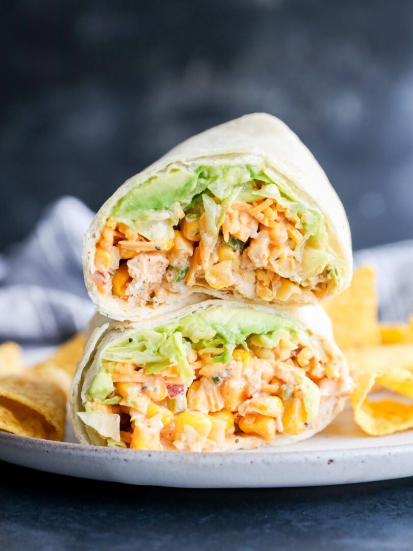 A close-up of a wrap cut in half, filled with lettuce, corn, diced vegetables, and a creamy filling, served on a plate with tortilla chips.