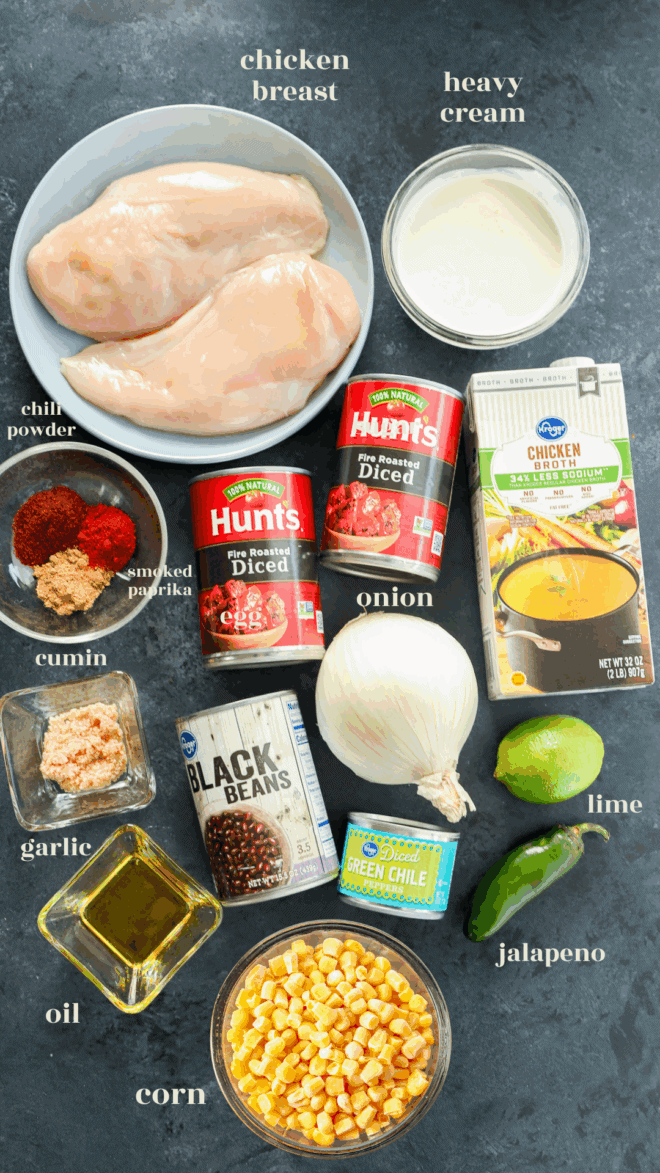 A flat lay of Creamy Chicken Tortilla Soup ingredients, featuring raw chicken breasts, heavy cream, broth, diced tomatoes, chili powder, smoked paprika, cumin, corn, black beans, onion, garlic, oil, lime, and jalapeño.