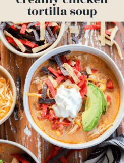 A bowl of creamy Chicken Tortilla Soup topped with sour cream, avocado slices, and colorful tortilla strips sits on a rustic wooden table, surrounded by bowls of shredded cheese and extra tortilla strips.