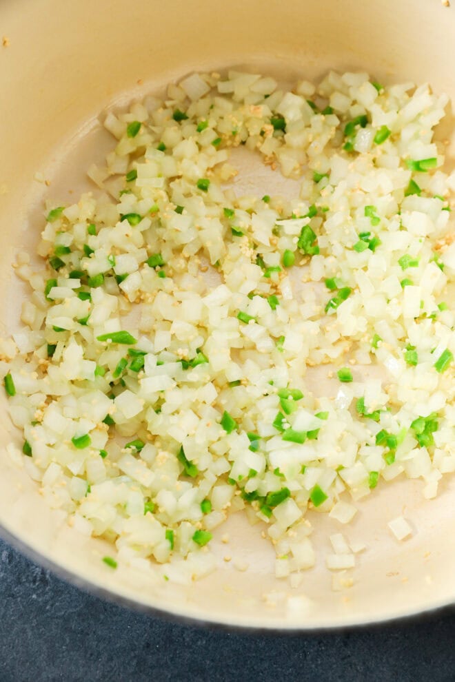 Diced onions, garlic, and green peppers are being sautéed in a light-colored pan.