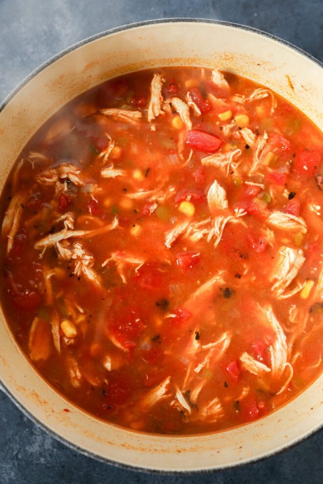 A pot filled with creamy chicken tortilla soup, featuring shredded chicken, corn, tomatoes, and herbs in a rich tomato-based broth. Steam rises from the hot, freshly cooked soup.