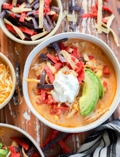 A bowl of stew topped with sliced avocado, sour cream, and colorful tortilla strips sits on a rustic wooden table, surrounded by bowls of shredded cheese and extra tortilla strips for the perfect finishing touch.