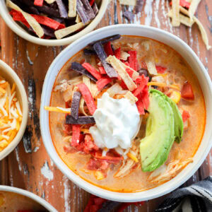 A bowl of stew topped with sliced avocado, sour cream, and colorful tortilla strips sits on a rustic wooden table, surrounded by bowls of shredded cheese and extra tortilla strips for the perfect finishing touch.