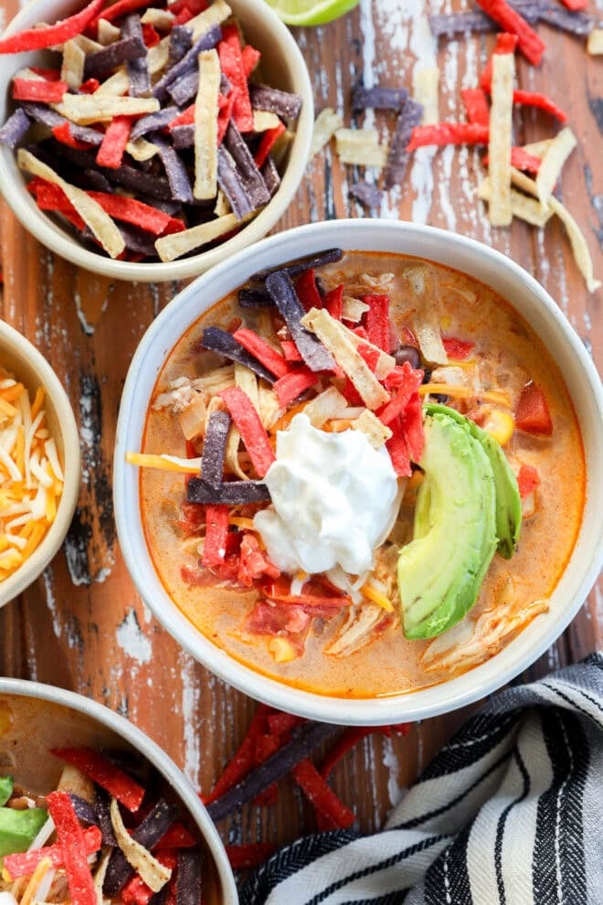 A bowl of stew topped with sliced avocado, sour cream, and colorful tortilla strips sits on a rustic wooden table, surrounded by bowls of shredded cheese and extra tortilla strips for the perfect finishing touch.