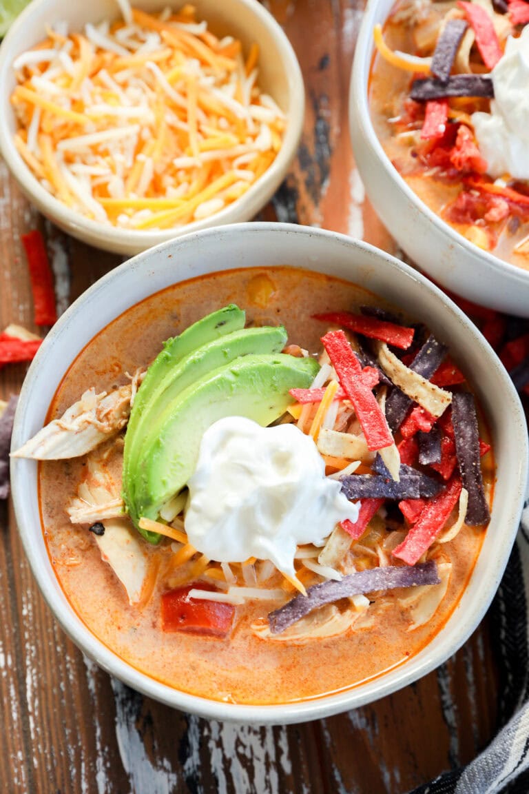 A bowl of Creamy Chicken Tortilla Soup topped with sliced avocado, shredded cheese, sour cream, and colorful tortilla strips sits on a rustic wooden table next to another bowl of soup and a small bowl of shredded cheese.