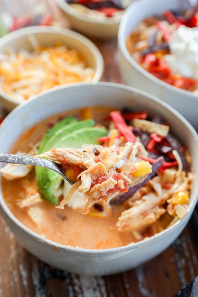 A close-up of a bowl of Creamy Chicken Tortilla Soup topped with sliced avocado, shredded cheese, and colorful tortilla strips, with a spoonful of this hearty soup being lifted in the foreground.
