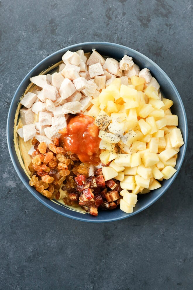 A bowl of curried chicken salad filled with chopped cooked chicken, diced pineapple, diced apples, golden raisins, chopped dates, chutney, and black pepper sits on a dark gray surface.