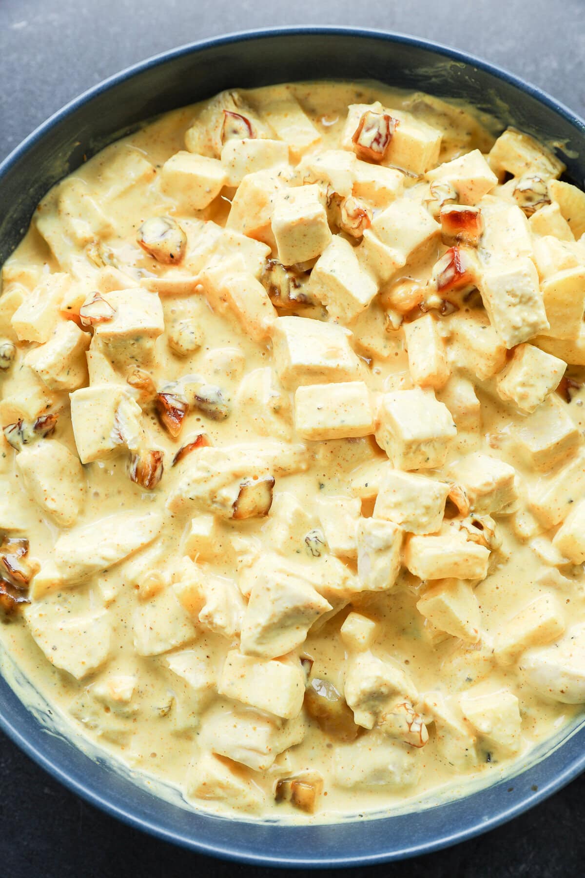 A close-up of a creamy yellow curry dish with cubed chicken, raisins, and fruit, all coated in a rich, spiced sauce for curried chicken salad, served in a dark bowl.