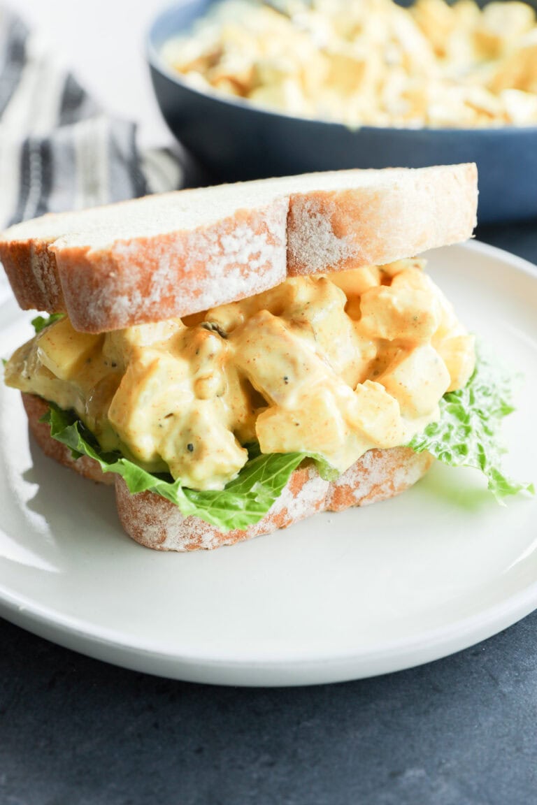 A close-up of an curried chicken salad sandwich with leafy lettuce on white bread, served on a white plate. In the background, a bowl of salad sits beside a dish of curried chicken salad for added variety.