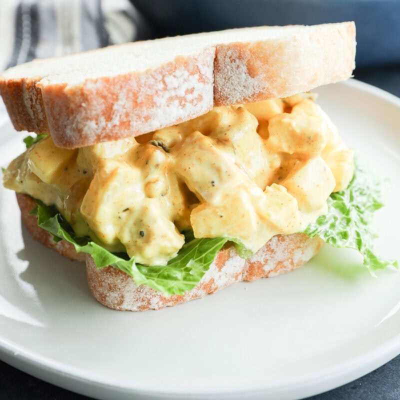 A close-up of an curried chicken salad sandwich with leafy lettuce on white bread, served on a white plate. In the background, a bowl of salad sits beside a dish of curried chicken salad for added variety.